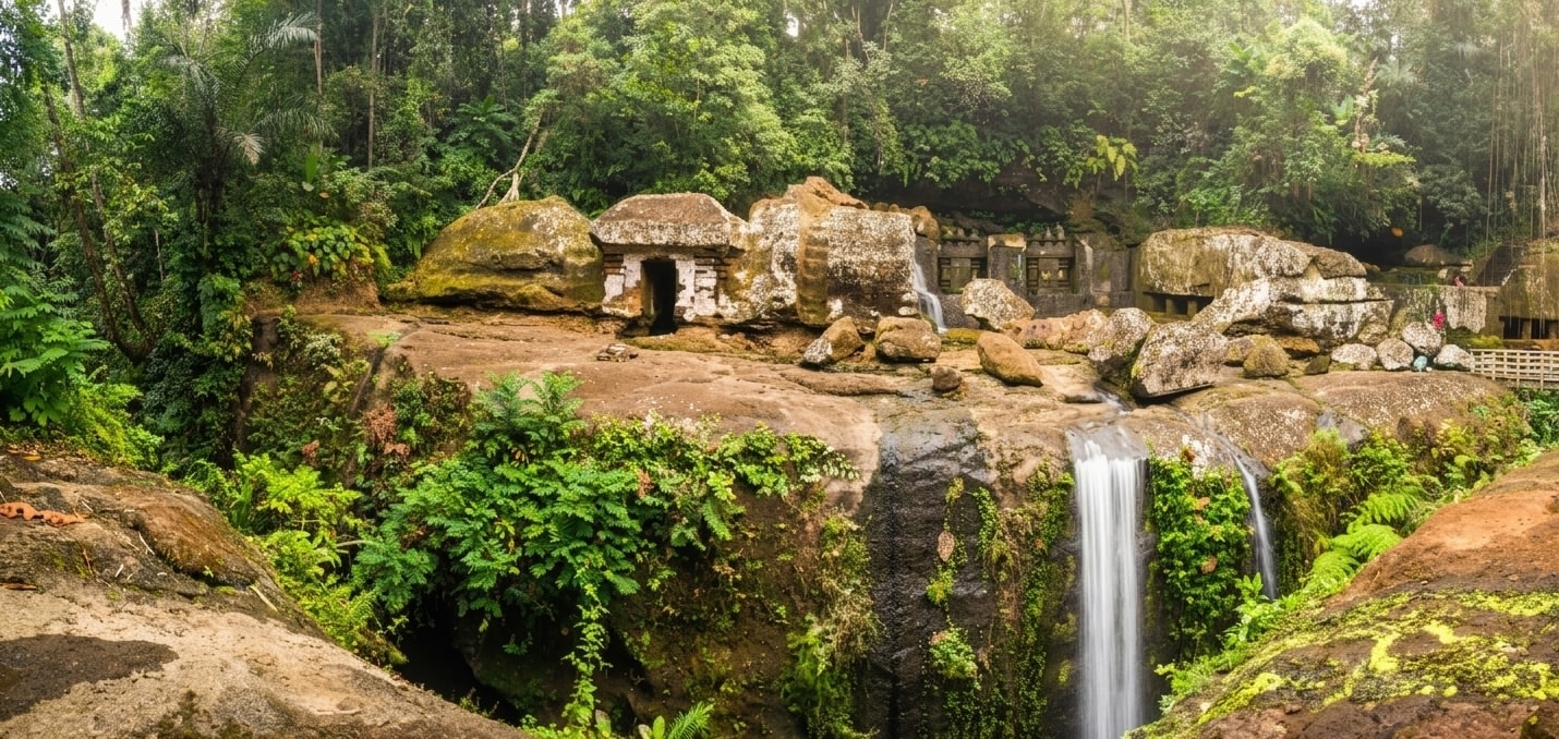 A panoramic view of the ancient Gunung Kawi temple complex in Tampaksiring, Bali, Indonesia, set within a lush tropical rainforest. The image captures a series of large, rock-cut shrines, or candi, carved directly into a massive cliff face on the left. A smaller, built temple structure sits on a ledge nearby. To the right, a dramatic waterfall cascades down the rock face into a river gorge. Visitors can be seen walking on a wooden bridge and stairs, which lead through the verdant landscape and connect different areas of the sacred site.