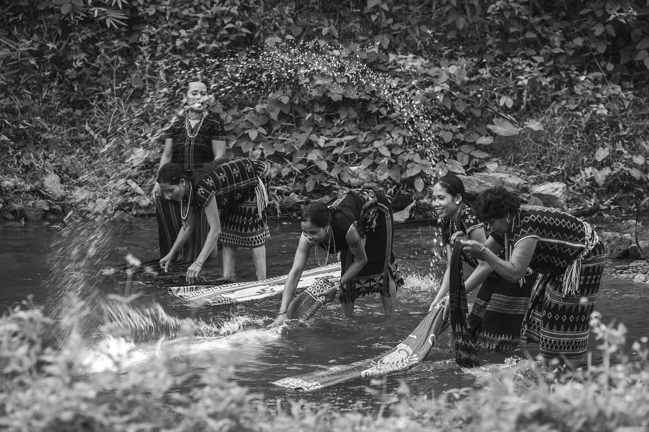 A black and white action photograph of four Katu women washing large woven textiles in a splashing river, surrounded by rocks and foliage in Vietnam.
