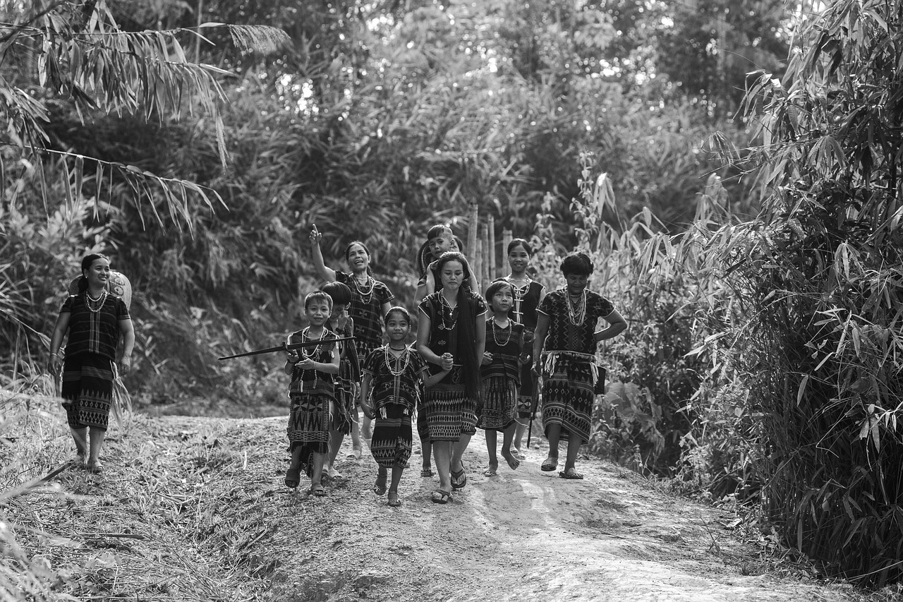 A black and white photograph of a group of Katu women and children wearing traditional patterned clothing walking in a line on a dirt path through a dense forest in Vietnam.