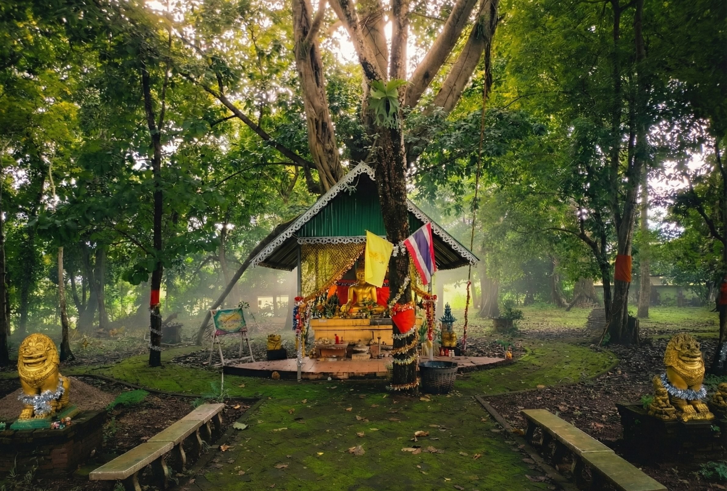 A small, decorated Buddhist shrine with a green corrugated roof sits in a lush, green forest. It features a golden Buddha statue, colorful flags, and offerings, with two golden lion (Singha) guardian statues flanking a mossy brick pathway leading up to it.