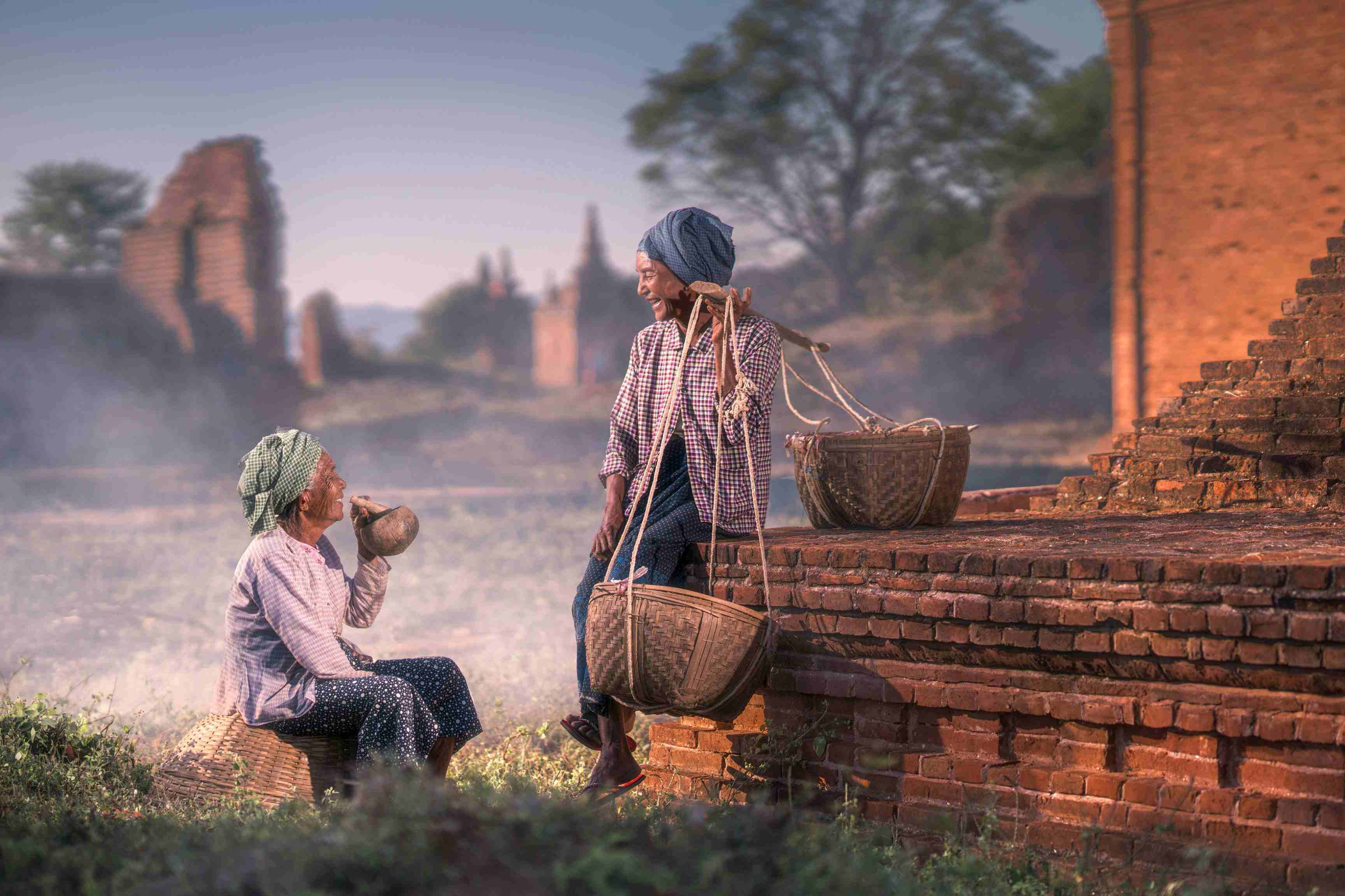 Two elderly local women resting on ancient brick ruins in Bagan, Myanmar; one smokes a traditional cheroot pipe while the other sits beside woven baskets, set against a backdrop of misty historical pagodas.