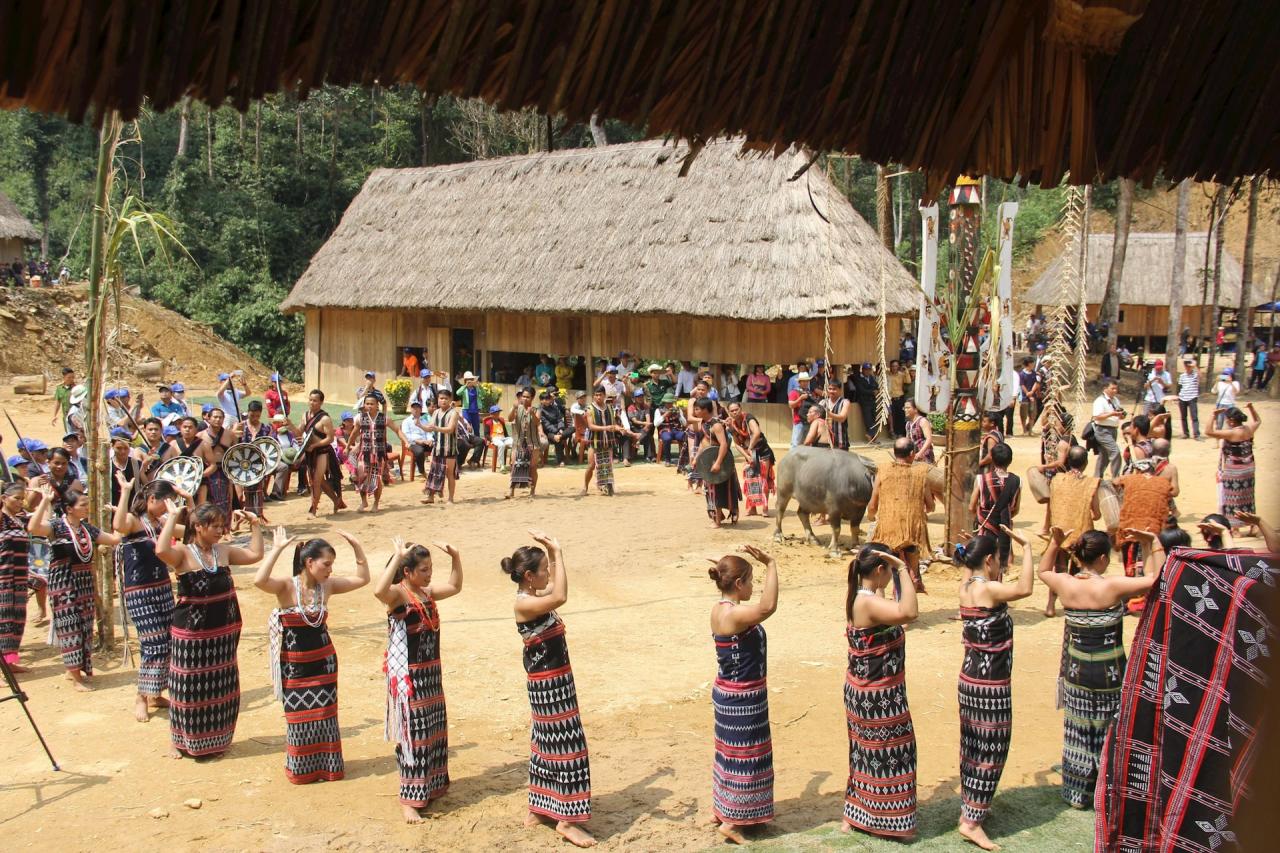 A group of Co Tu villagers in traditional black and red woven clothing perform a ritual dance in a circle around a water buffalo and a decorated pole, with a large thatched-roof stilt house in the background.