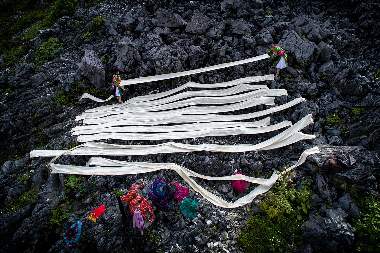 A candid ethnographic photograph of a large Hmong community gathering outdoors in a [village/marketplace] setting in [Southeast Asia/Northern Vietnam/Laos]. Numerous individuals of different generations mingle, with many women wearing distinctive traditional Hmong embroidered attire, silver jewellery, and headdresses. The atmosphere appears engaged and communal.
